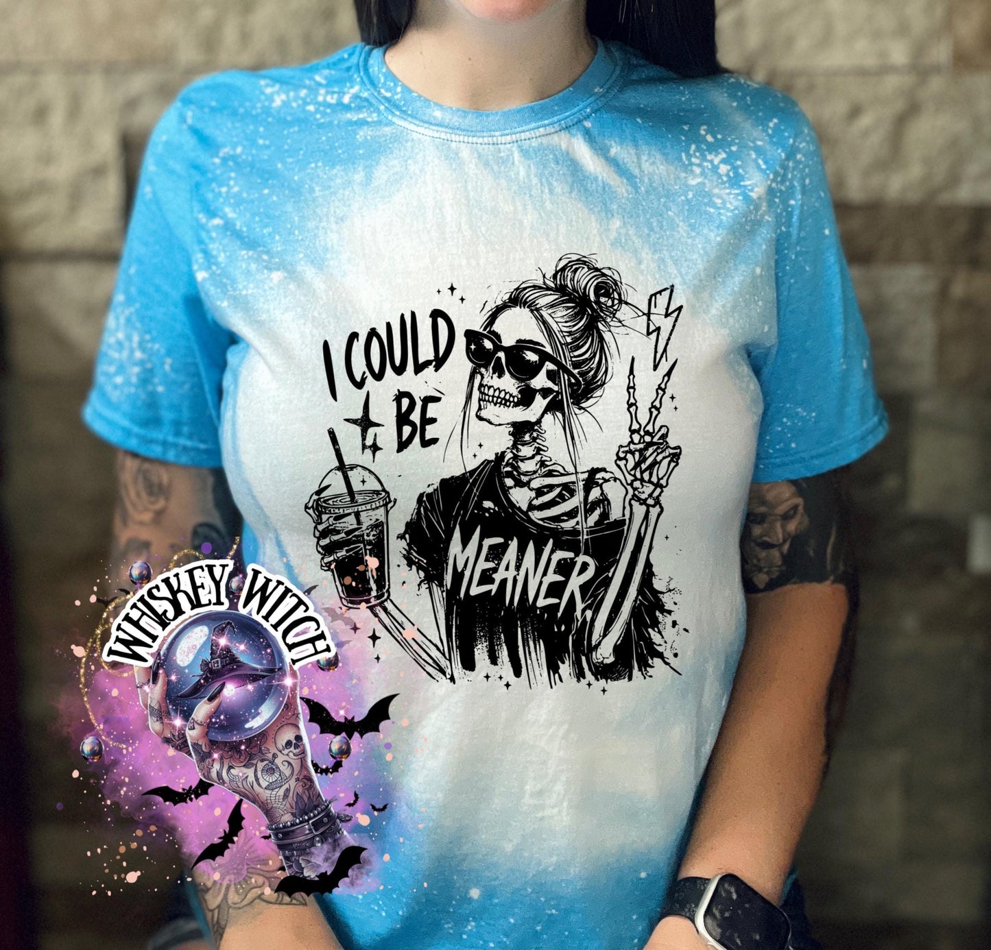 A saphire blue t-shirt with bleach white splatter effect with a graphic of a skeleton wearing sunglasses, holding a drink, and making a peace sign. The text on the shirt reads "I COULD BE MEANER.”