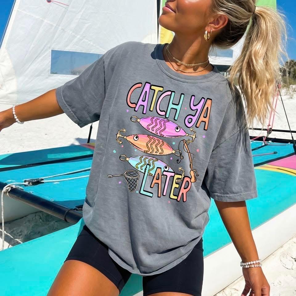 Cute lake day graphic tee with Catch Ya Later text, fishing lures, and a small fishing net design.