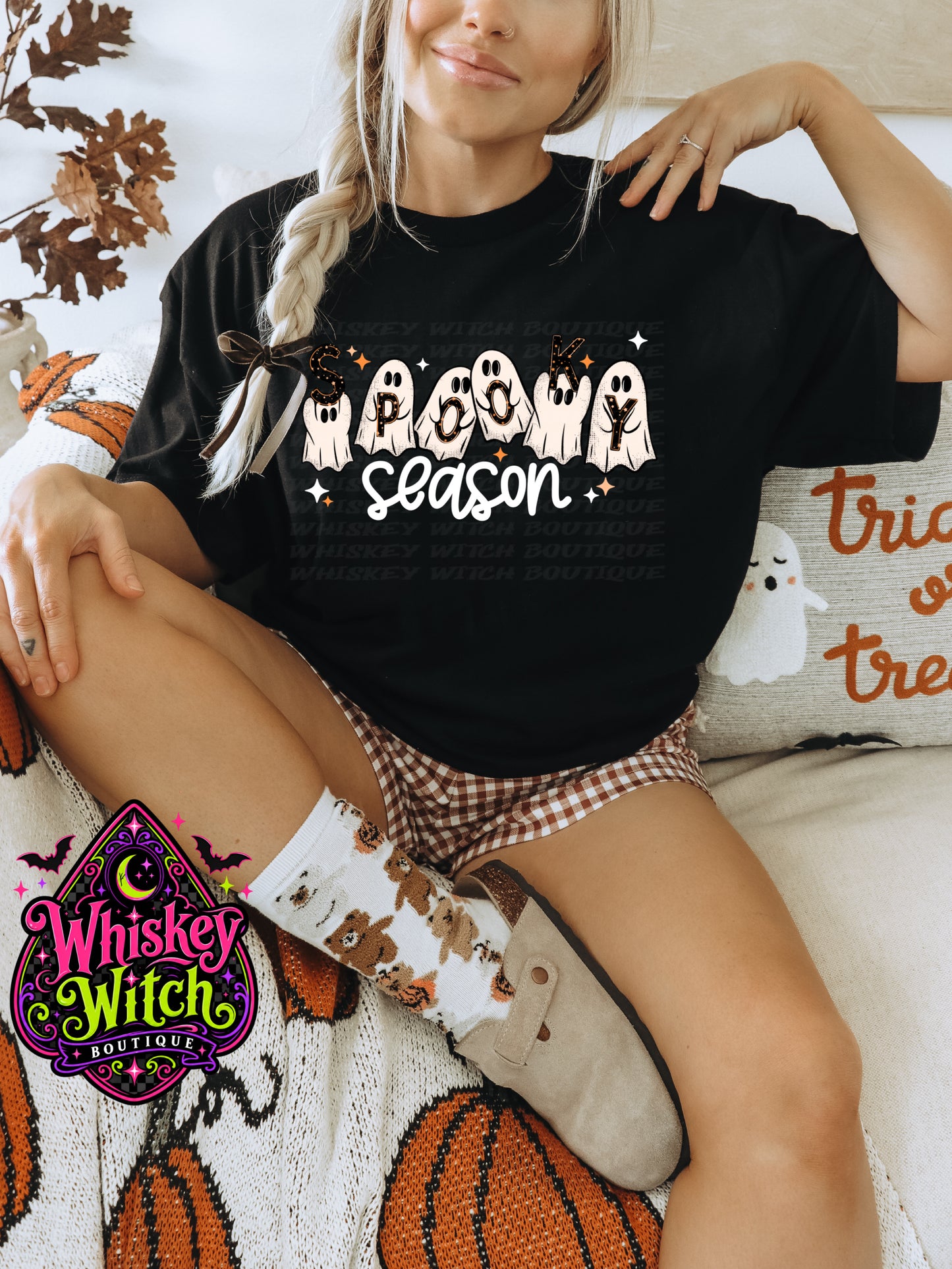 A woman wearing a black graphic t-shirt that reads 'Halloween season' with white ghosts and orange stars, paired with brown and white gingham shorts and white socks with a witch pattern. She is sitting on a cozy bed with autumn-themed pillows and blankets.