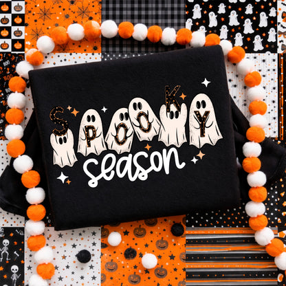 Woman wearing a black Spooky Season Halloween graphic T-shirt featuring cute white ghosts, orange stars and retro lettering.
