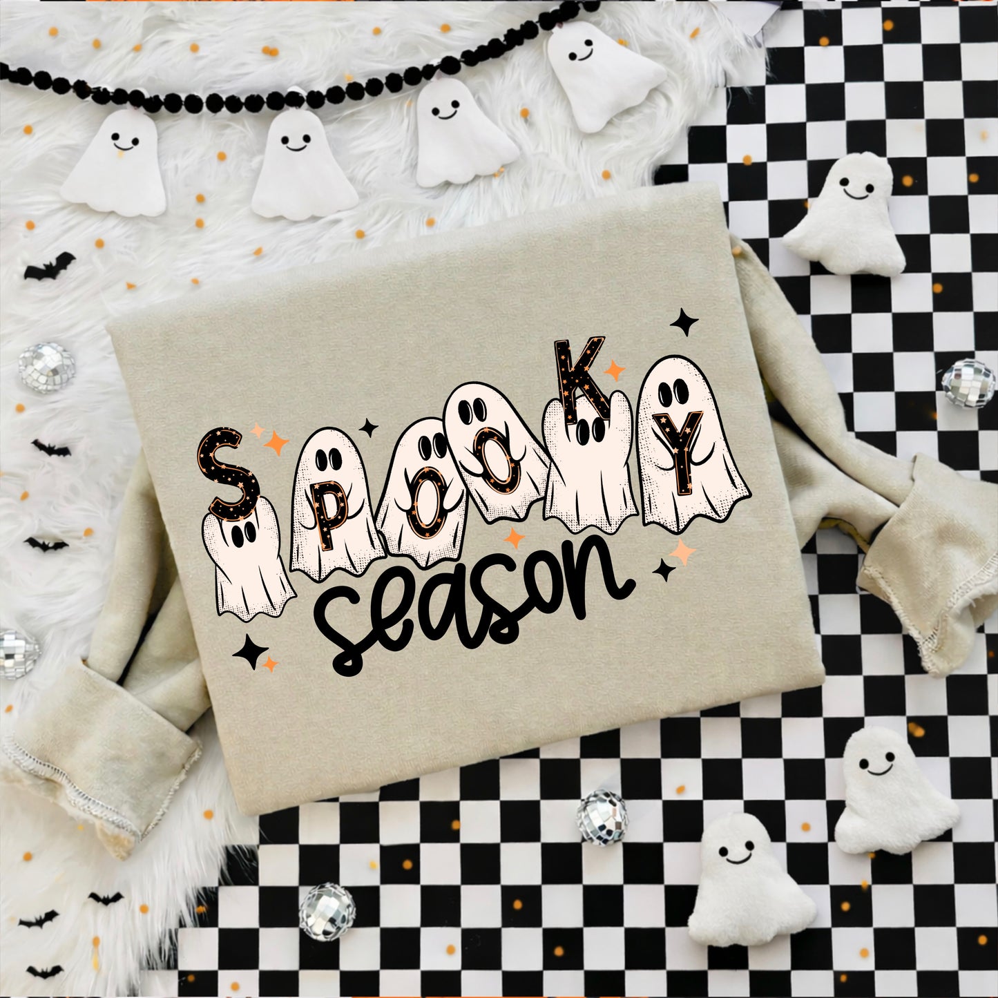 A beige sweatshirt laid flat with a 'Spooky Season' graphic featuring ghost illustrations and black lettering, styled with Halloween decorations including white ghost cutouts, a black-and-white checkered mat, fluffy white fabric, and small black bat charms.