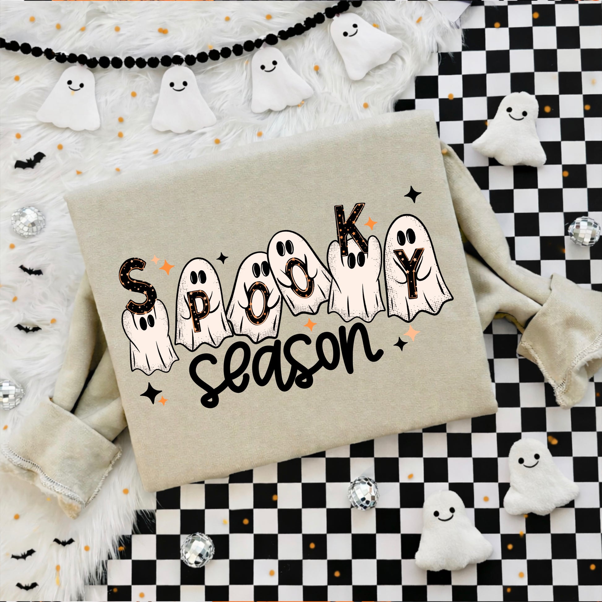 A beige sweatshirt laid flat with a 'Spooky Season' graphic featuring ghost illustrations and black lettering, styled with Halloween decorations including white ghost cutouts, a black-and-white checkered mat, fluffy white fabric, and small black bat charms.