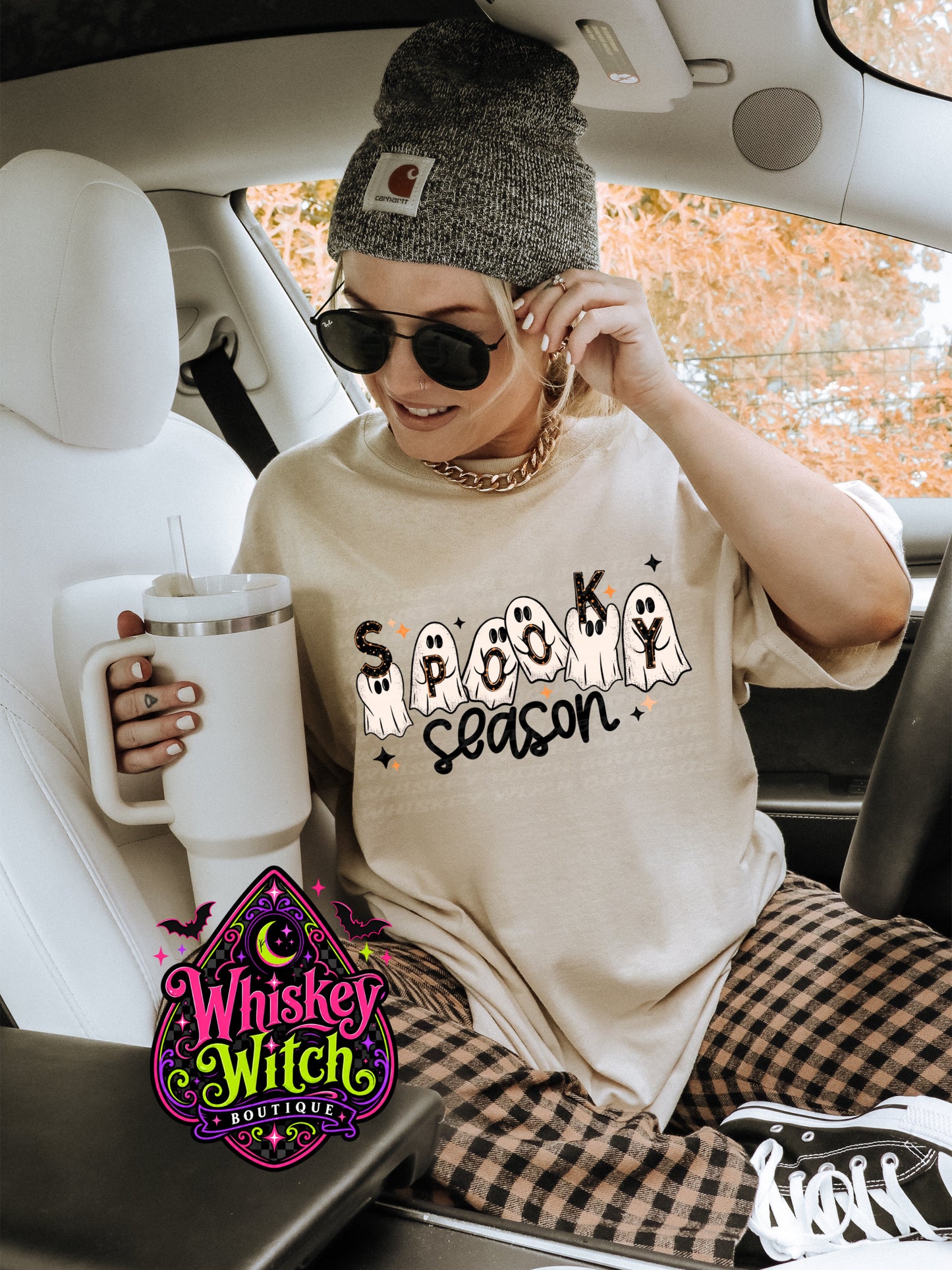 A woman sitting in a car wearing a beige graphic t-shirt that says 'SPOOK season' with ghost illustrations, a gray knit beanie, sunglasses, and plaid pants. She is holding a white insulated tumbler.