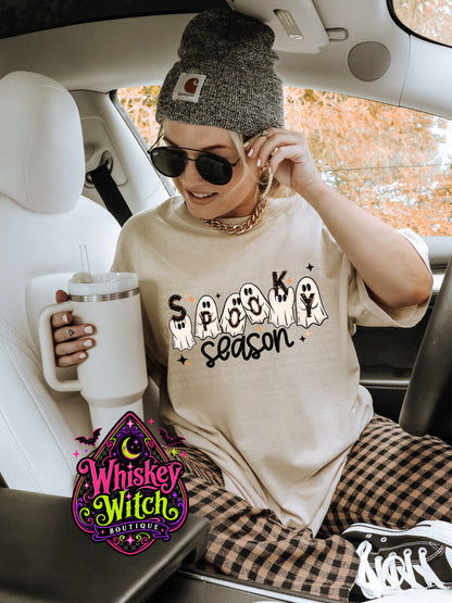 A woman sitting in a car wearing a beige graphic t-shirt that says 'SPOOK season' with ghost illustrations, a gray knit beanie, sunglasses, and plaid pants. She is holding a white insulated tumbler.