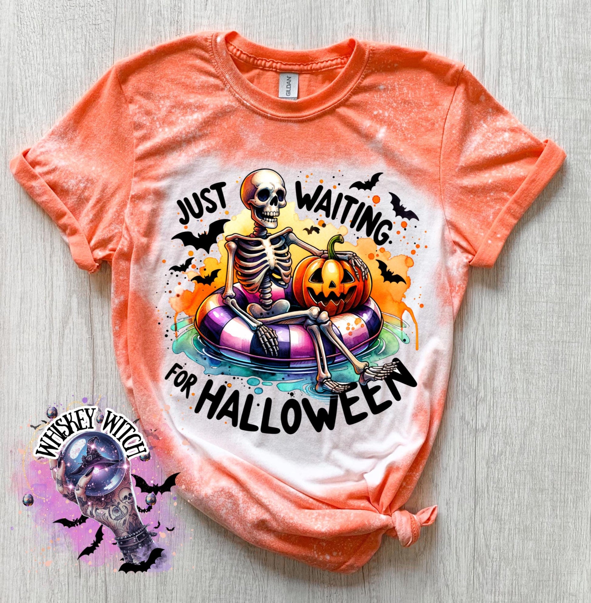 Orange t-shirt with a bleached effect featuring a skeleton sitting in a pool float with a pumpkin. The text on the shirt reads "Just Waiting for Halloween".
