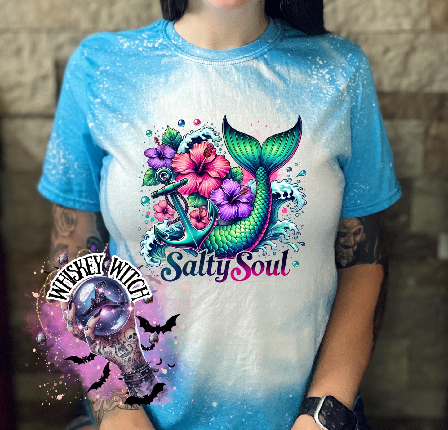 A sapphire blue   t-shirt with a bleach splatter effect. The shirt features a colorful graphic of a mermaid tail, anchor, flowers, and the text "Salty Soul". The mermaid tail is green and blue.