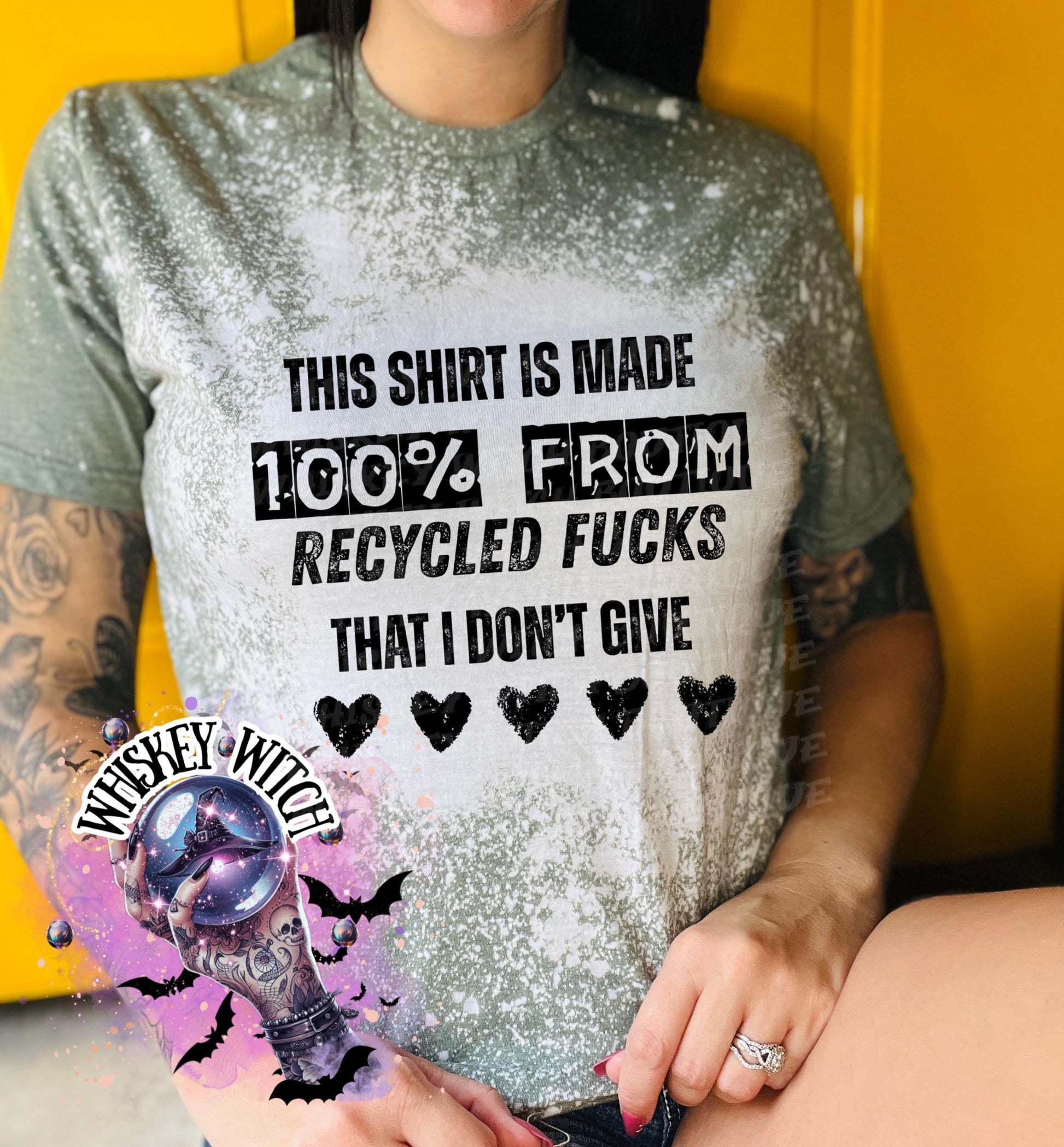 Military Green  t-shirt with the text "THIS SHIRT IS MADE 100% FROM RECYCLED FUCKS THAT I DON'T GIVE" in black. The shirt has a bleached effect and four black heart graphics. A pair of black and white checkered sneakers are at the bottom.