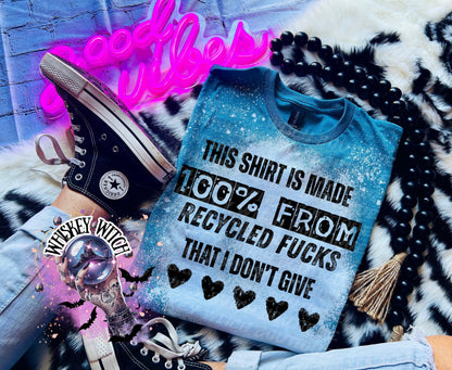 A Galapagos blue t-shirt with the text "THIS SHIRT IS MADE 100% FROM RECYCLED FUCKS THAT I DON'T GIVE" in black. The shirt has a bleached effect and four black heart graphics. A black Converse sneaker is visible.