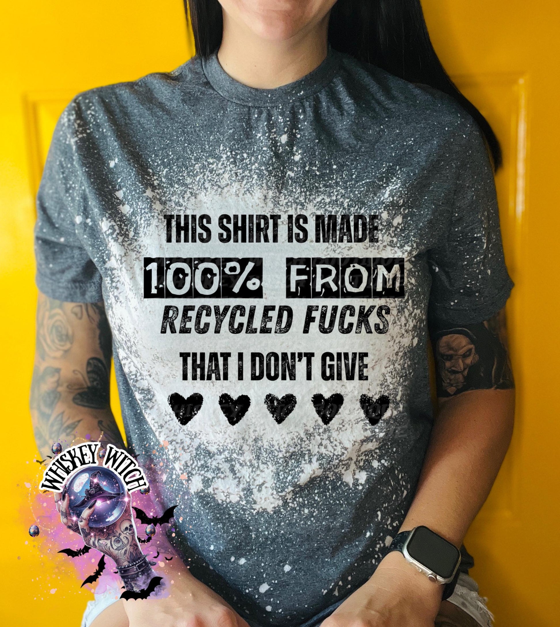 Gray t-shirt with the text "THIS SHIRT IS MADE 100% FROM RECYCLED FUCKS THAT I DON'T GIVE" in black. The shirt has a bleached effect and four black heart graphics. A pair of black and white checkered sneakers are at the bottom.
