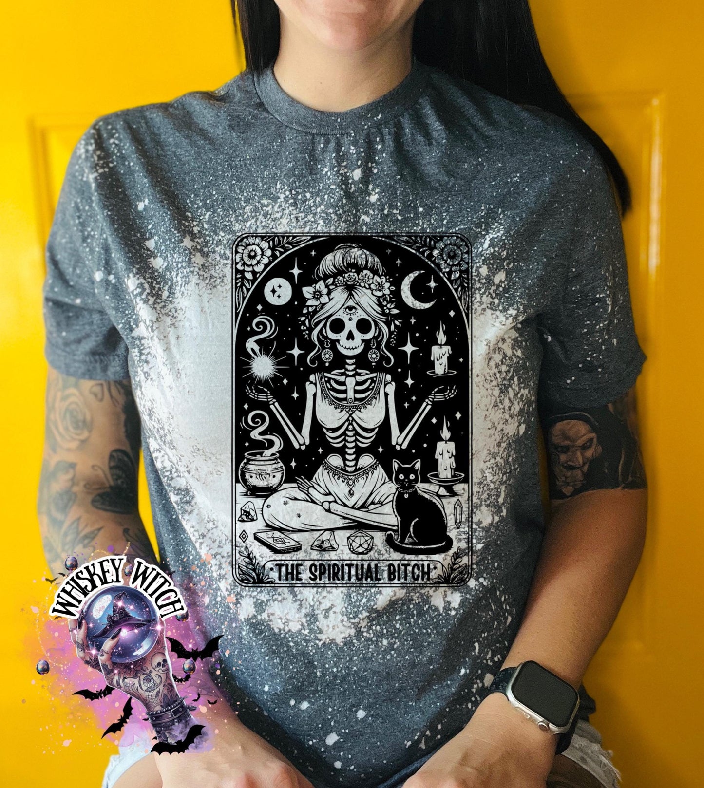 A bleached grey t-shirt with a black and white illustration of a skeleton woman with a cat and the text "The Spiritual Bitch".