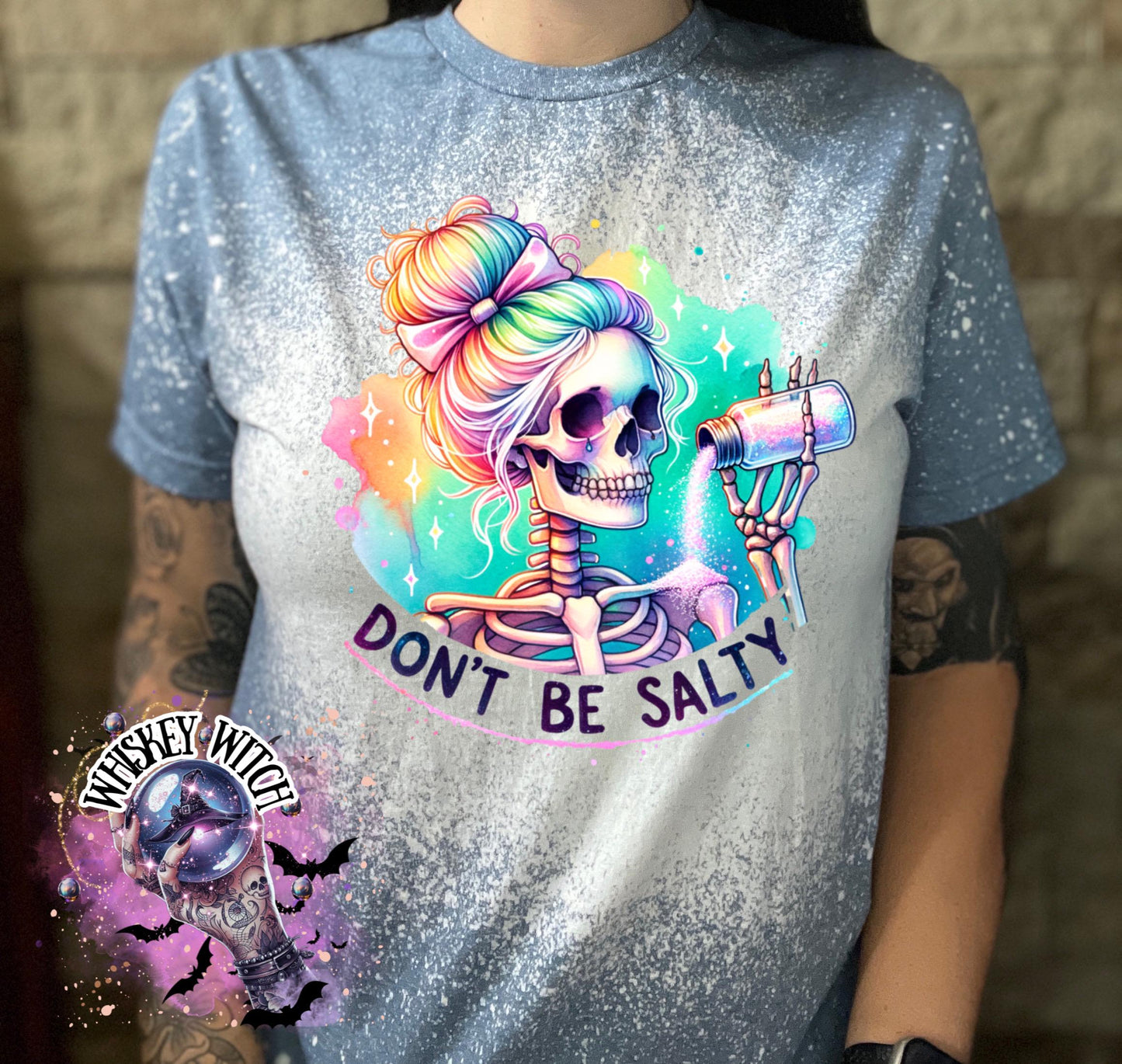 A indigo blue  t-shirt with a bleached effect featuring a skeleton with rainbow hair pouring salt from a shaker. The text "Don't Be Salty" is printed on the shirt