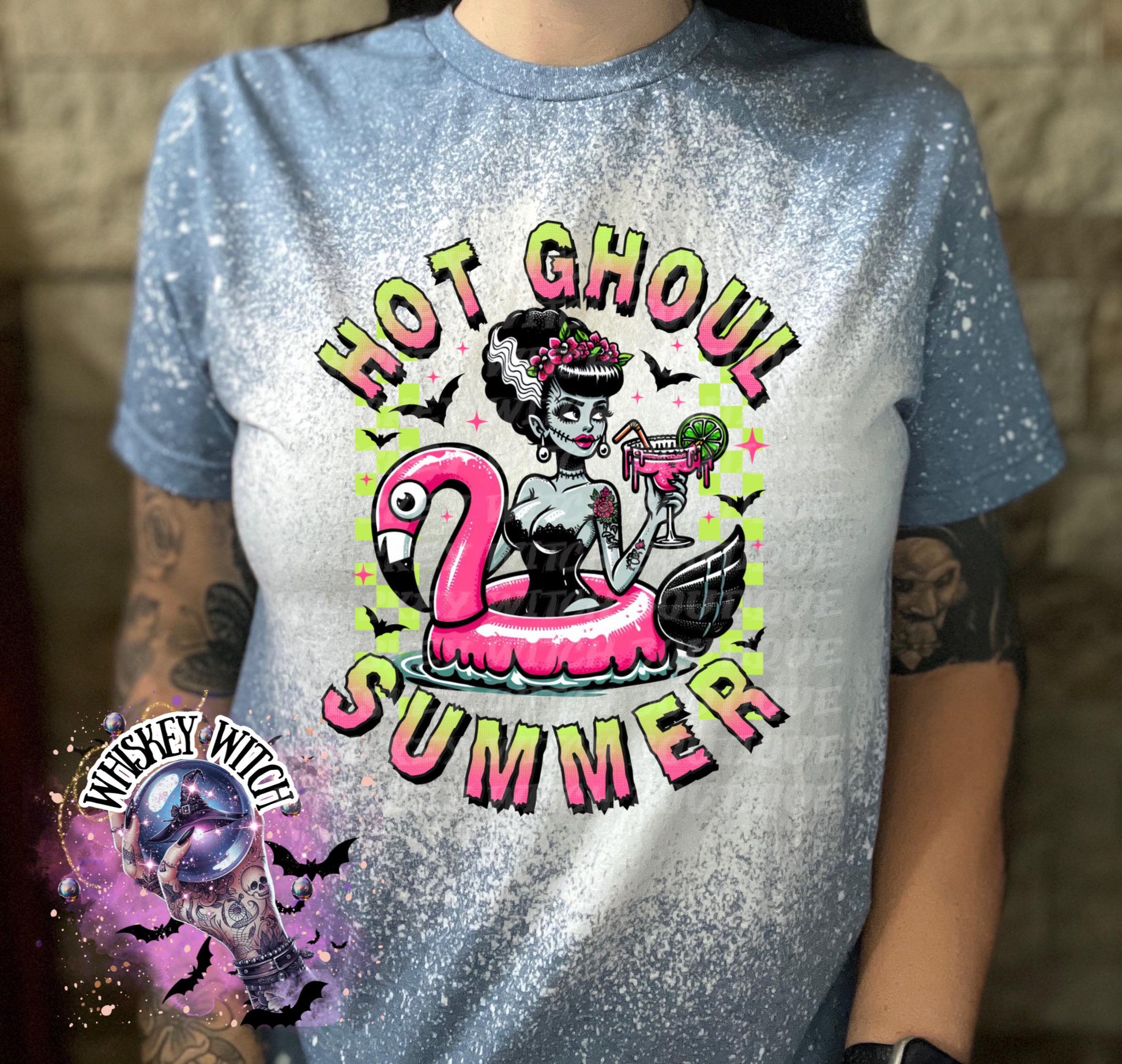 A indigo blue  and bleach splattered effect  t-shirt with a graphic of a zombie woman in a flamingo pool float, holding a cocktail. The text "Hot Ghoul Summer" is printed on the shirt.