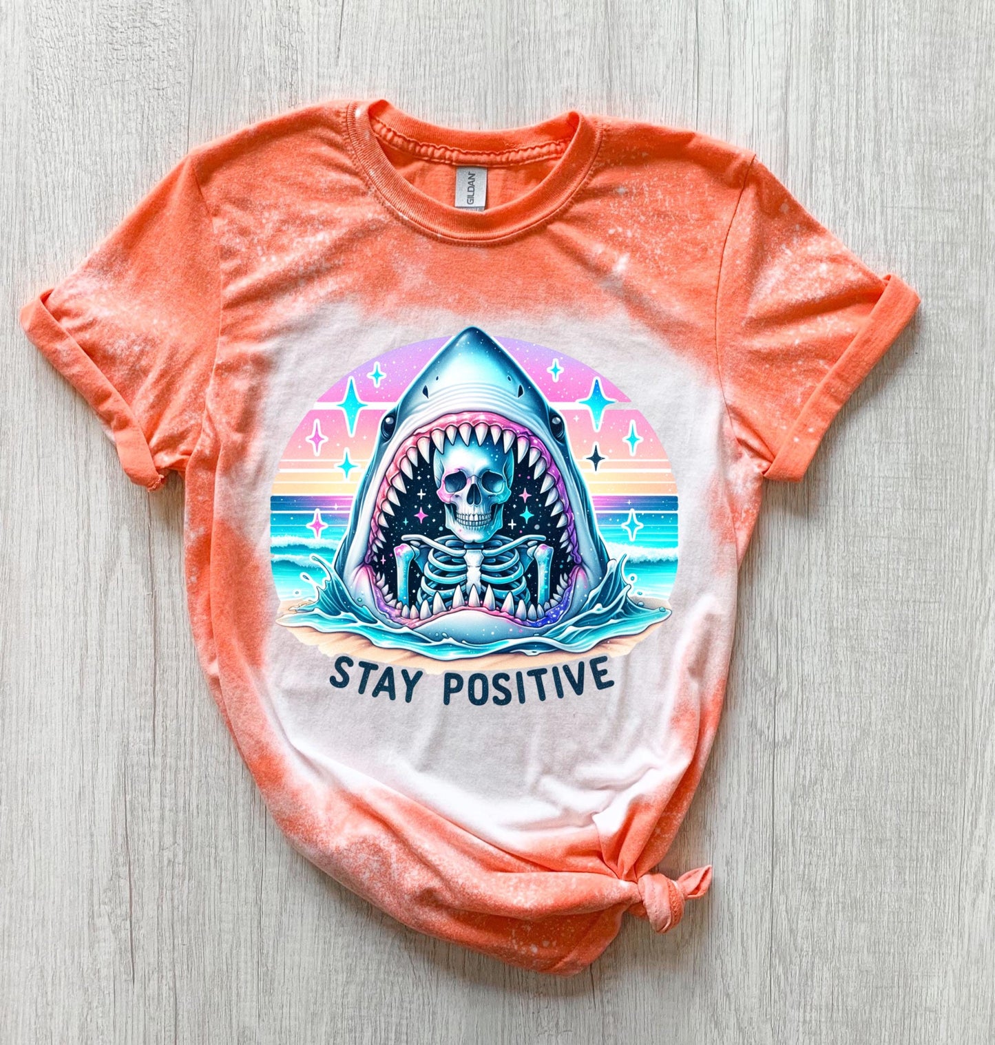 A Orange  t-shirt with a bleached design of a skeleton inside a shark's mouth. The shark is surrounded by a rainbow and stars. The text "Stay Positive" is below the shark.