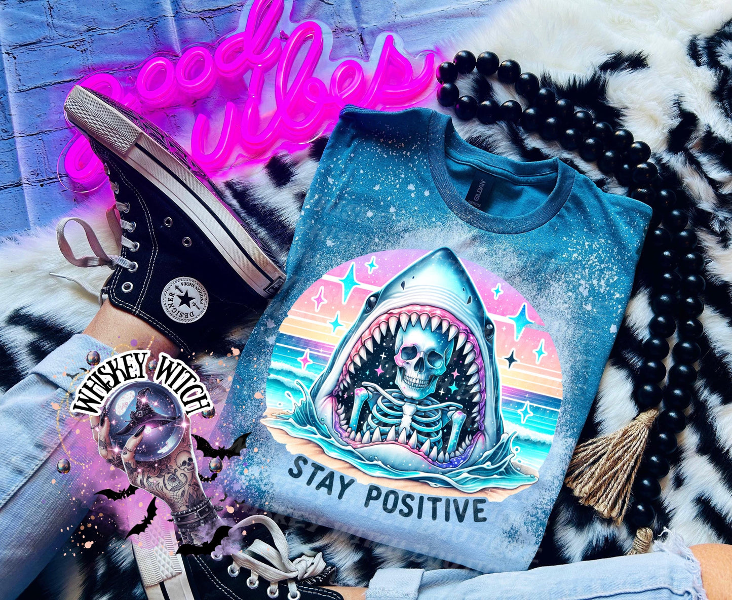 A Galapagos blue t-shirt with a bleached design of a skeleton inside a shark's mouth. The shark is surrounded by a rainbow and stars. The text "Stay Positive" is below the shark.