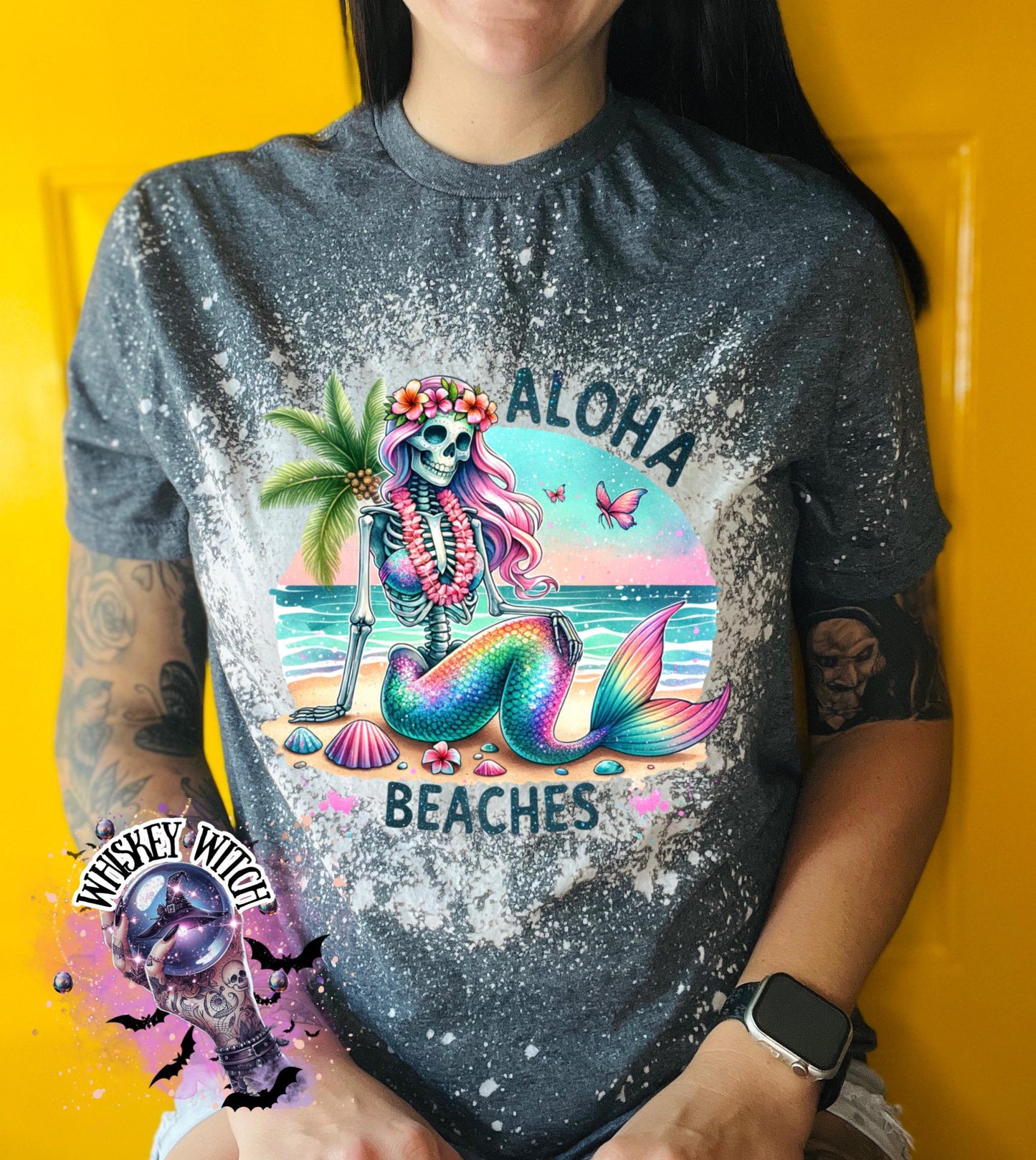 A grey t-shirt with a bleached effect features a colorful skeleton mermaid with a flower crown, palm tree, and seashells. The text "Aloha Beaches" is printed on the shirt.
