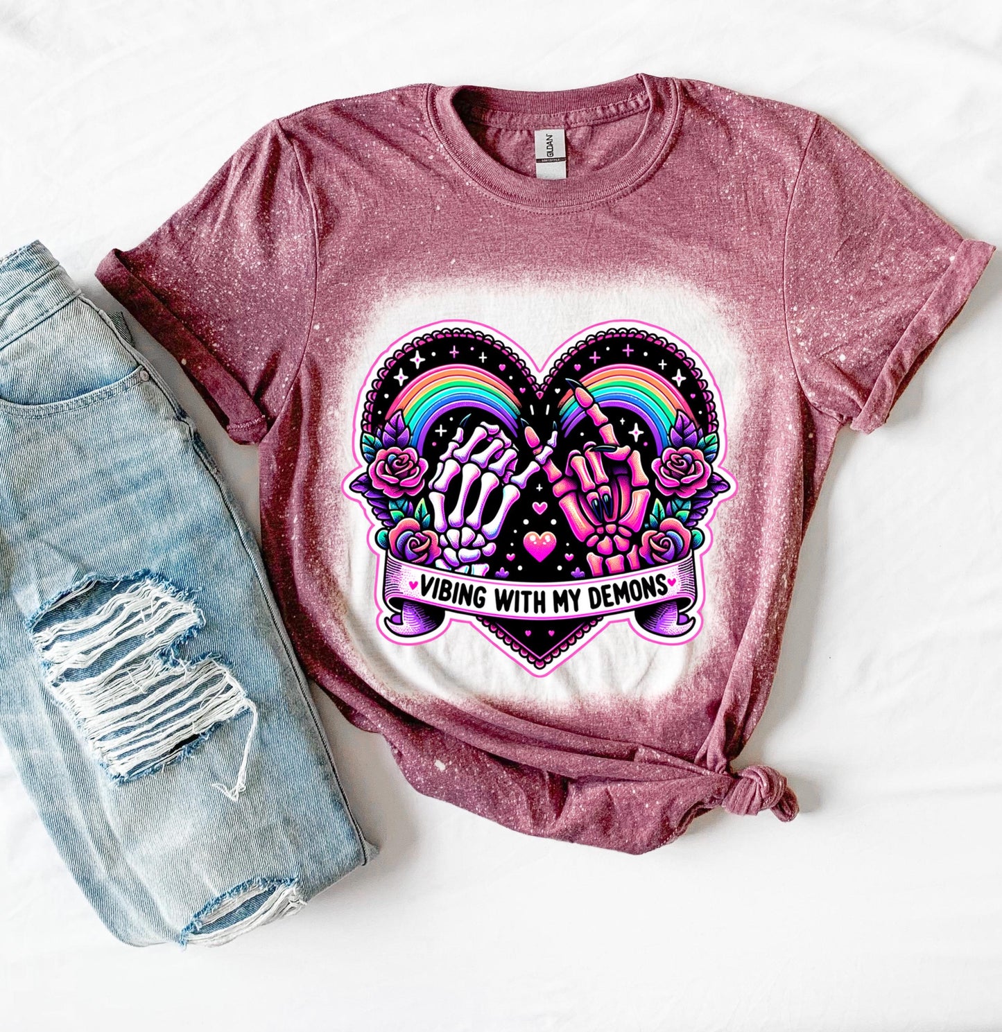A maroon t-shirt with a bleach-splattered design featuring a pink heart with a rainbow and two hands, one skeleton and one with claws, holding pinky fingers together. The text 'Vibing with my demons' is written below the heart.