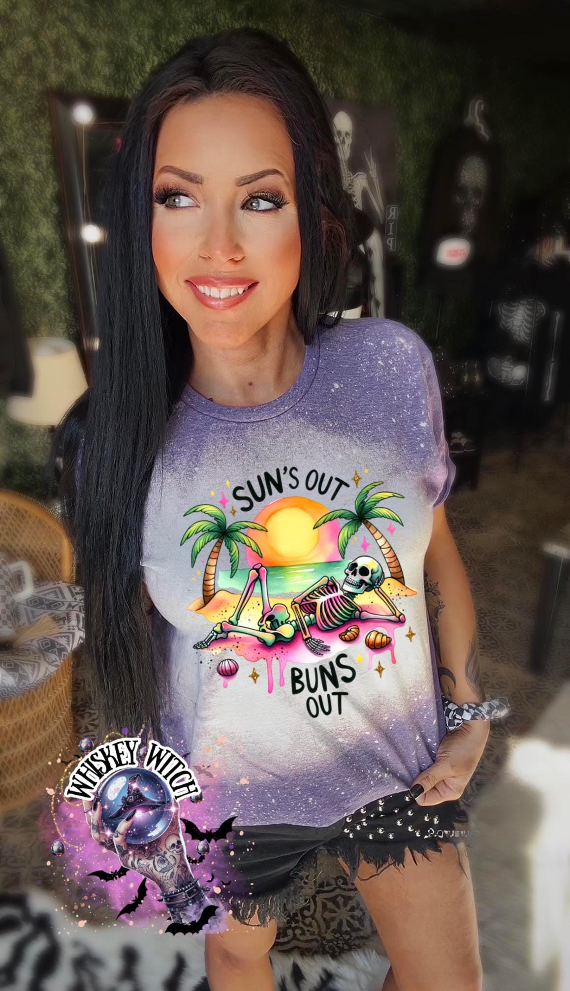 A purple  t-shirt with a graphic of a skeleton relaxing on a beach with palm trees and the text "Sun's Out Buns Out." The shirt has a bleached effect.