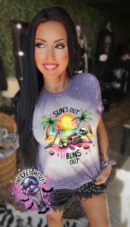 A purple  t-shirt with a graphic of a skeleton relaxing on a beach with palm trees and the text "Sun's Out Buns Out." The shirt has a bleached effect.