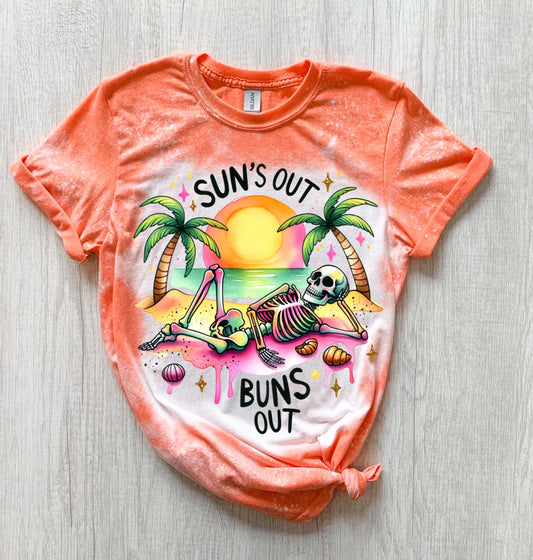 A Orange t-shirt with a graphic of a skeleton relaxing on a beach with palm trees and the text "Sun's Out Buns Out." The shirt has a bleached effect.