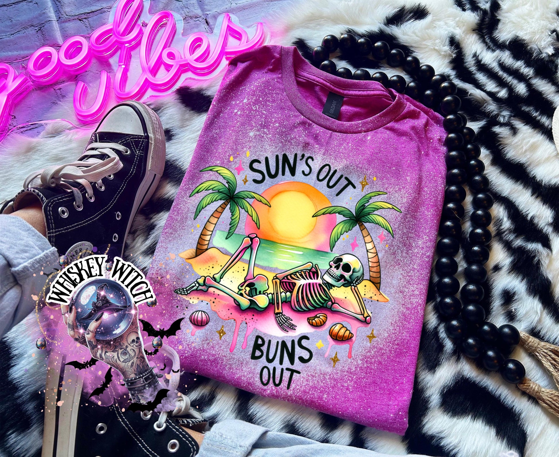 A Berry pink t-shirt with a graphic of a skeleton relaxing on a beach with palm trees and the text "Sun's Out Buns Out." The shirt has a bleached effect.