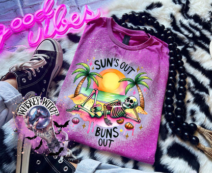 A Berry pink t-shirt with a graphic of a skeleton relaxing on a beach with palm trees and the text "Sun's Out Buns Out." The shirt has a bleached effect.