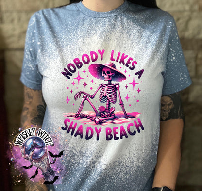 A indigo blue  t-shirt with a bleached design featuring a skeleton wearing a hat, the text "Nobody Likes a Shady Beach," and star graphics. The shirt is paired with black and white checkered sneakers.