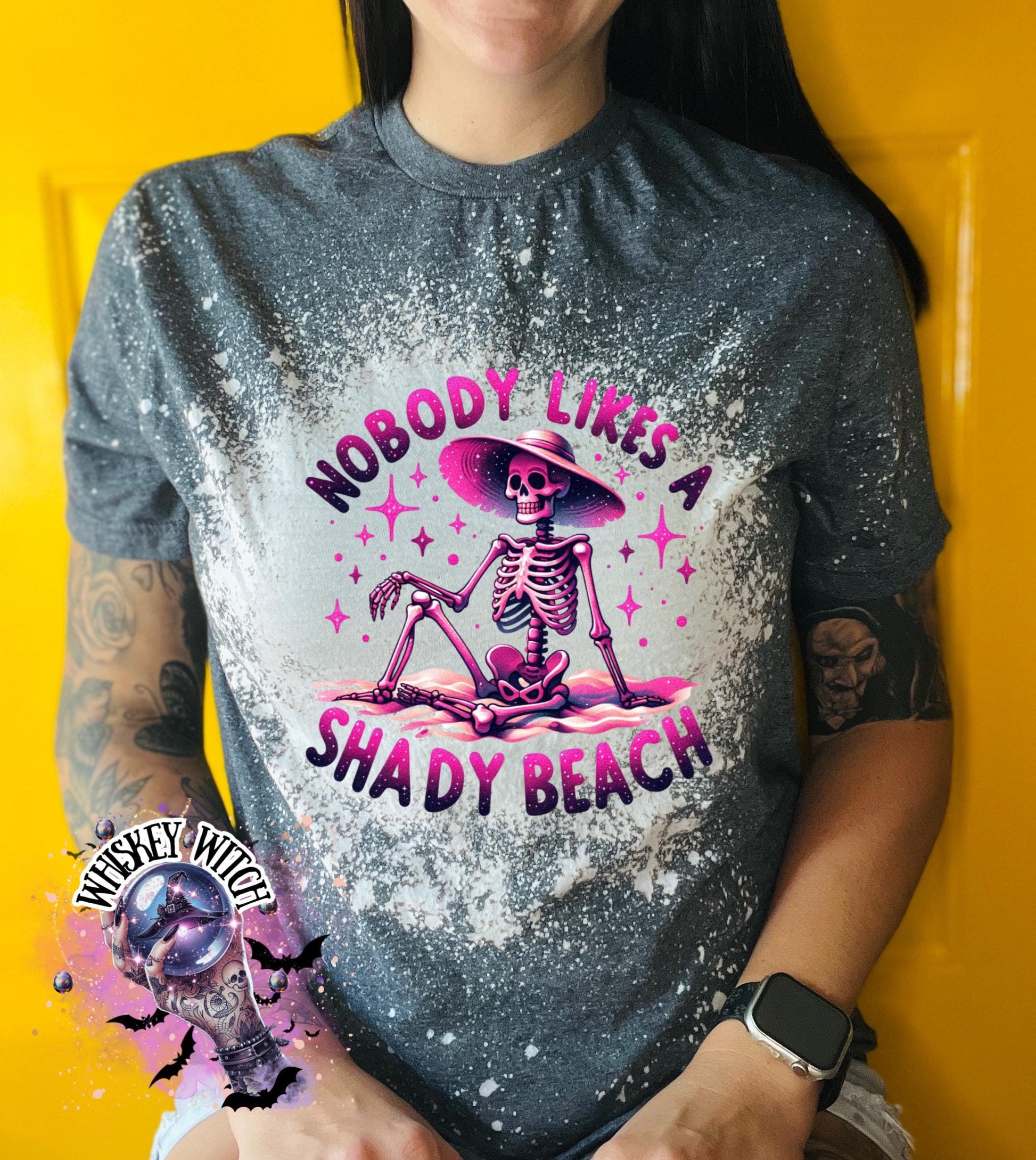 A dark grey  t-shirt with a bleached design featuring a skeleton wearing a hat, the text "Nobody Likes a Shady Beach," and star graphics. The shirt is paired with black and white checkered sneakers.