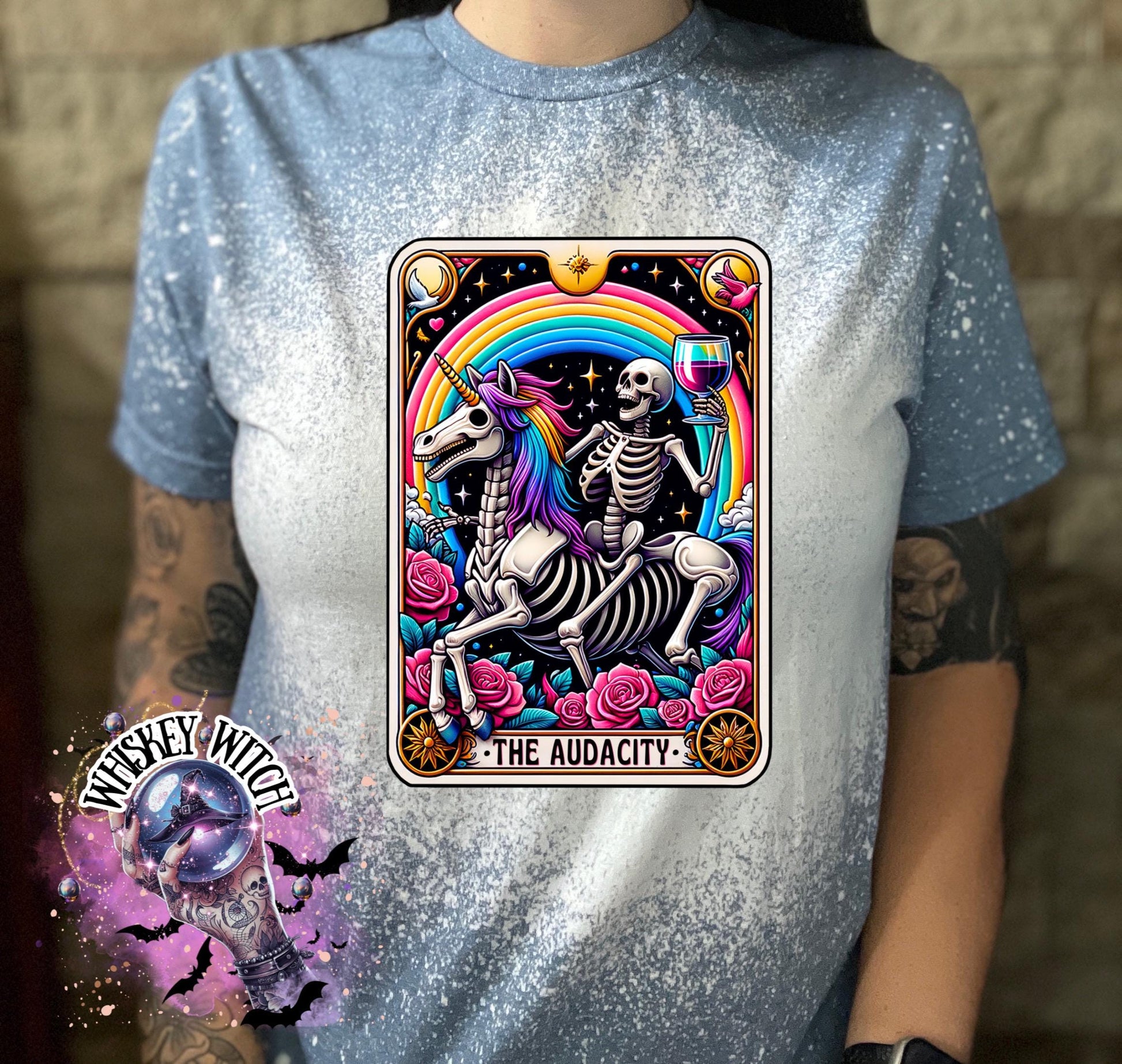 A indigo blue  t-shirt with a bleached design featuring a tarot card with the image of  skeleton holding a glass of wine  riding a unicorn with a rainbow behind them with the words of “The Audacity” at the bottom