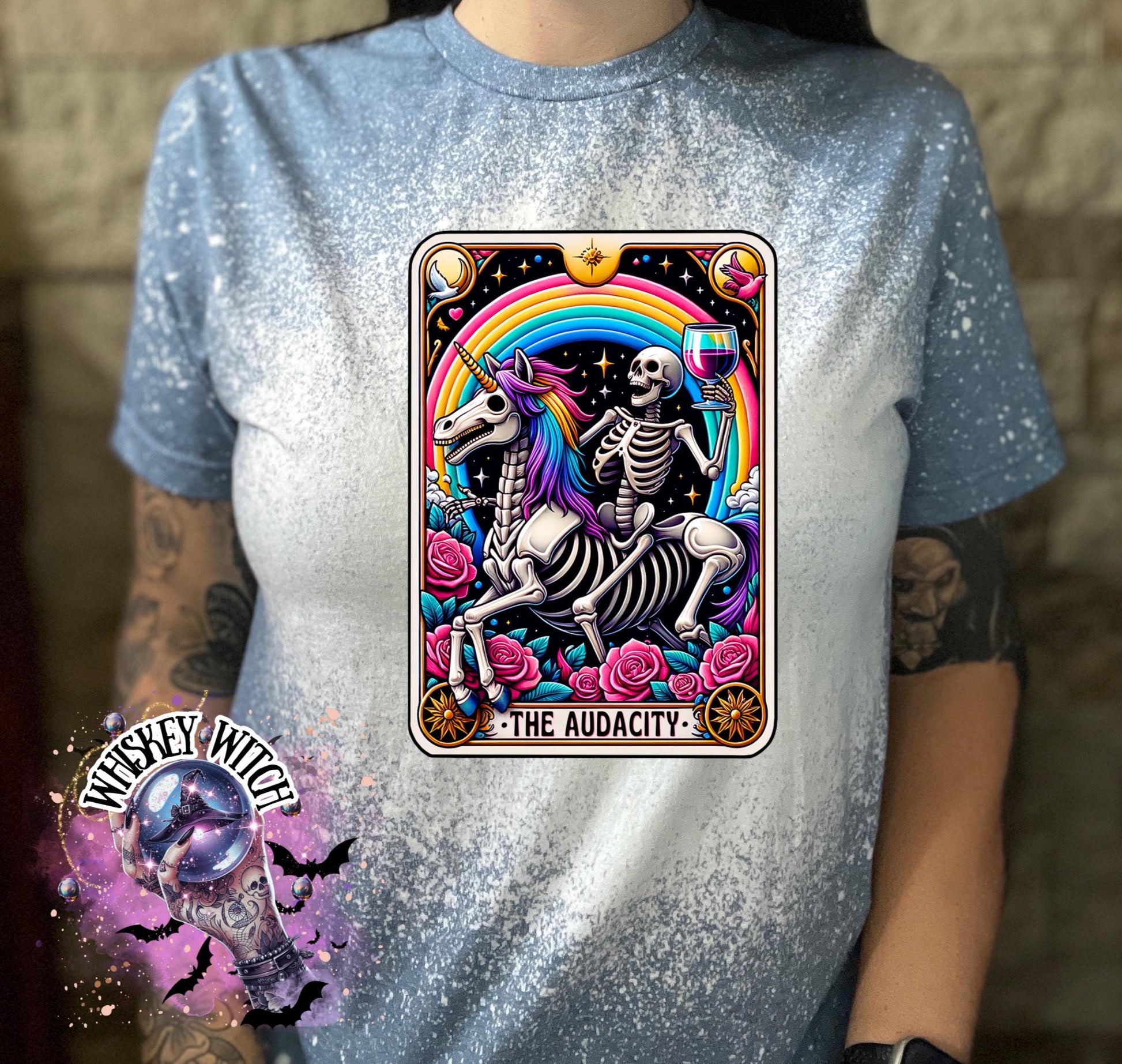 A Indigo blue   t-shirt with a bleached design featuring a tarot card with the image of  skeleton holding a glass of wine  riding a unicorn with a rainbow behind them with the words of “The Audacity” at the bottom