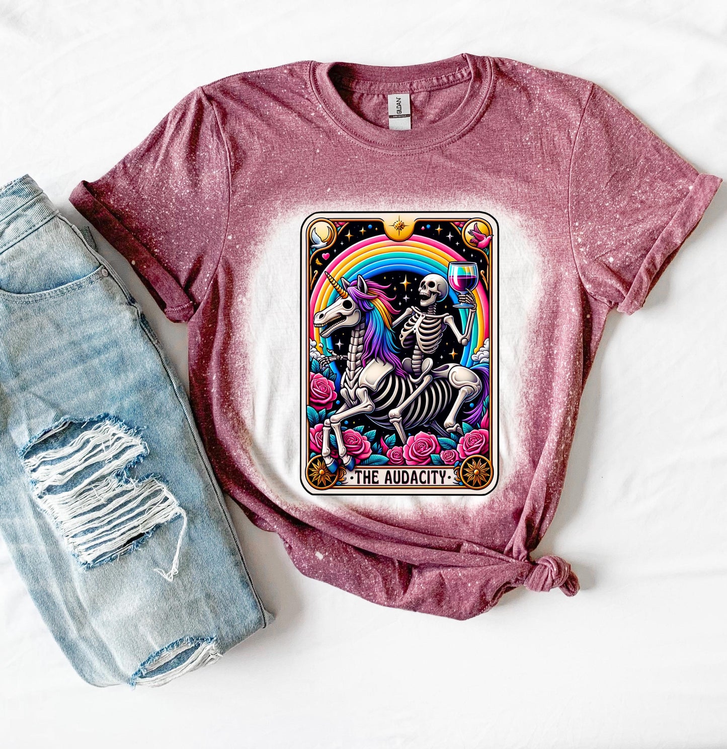 A maroon t-shirt with a bleached design featuring a tarot card with the image of  skeleton holding a glass of wine  riding a unicorn with a rainbow behind them with the words of “The Audacity” at the bottom