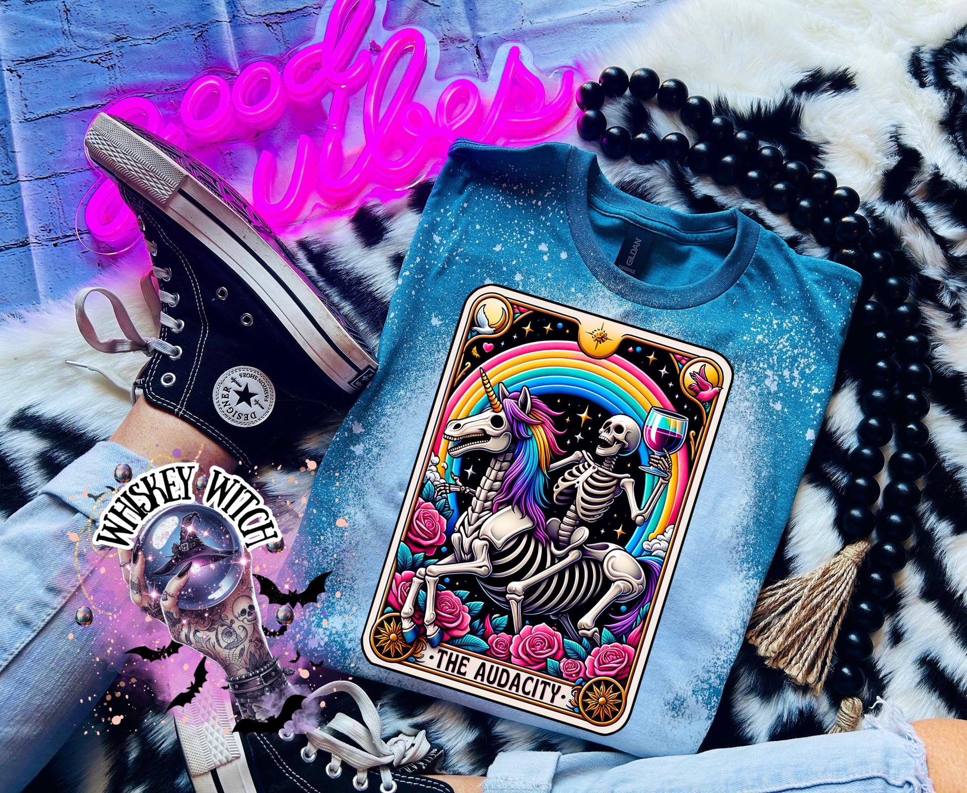 A Galapagos blue t-shirt with a bleached design featuring a tarot card with the image of  skeleton holding a glass of wine  riding a unicorn with a rainbow behind them with the words of “The Audacity” at the bottom