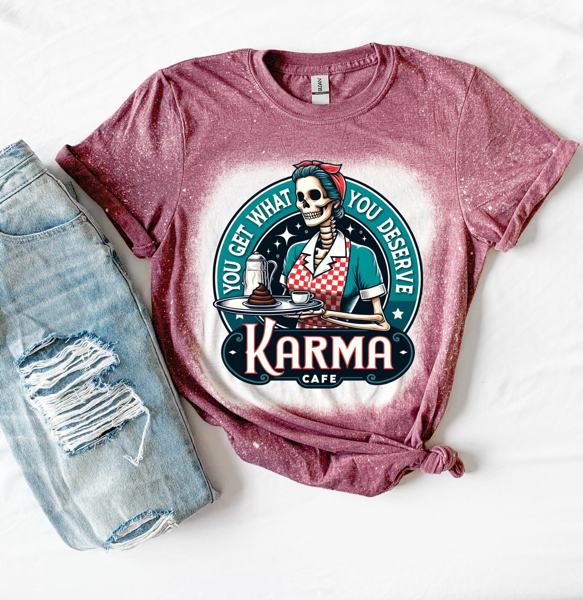 A maroon t-shirt with a bleached design featuring a skeleton wearing a red and white dress and holding a tray with a cup of coffee and a piece of cake. The text "You get what you deserve" is written around the skeleton. Below the skeleton is the text "Karma Cafe".