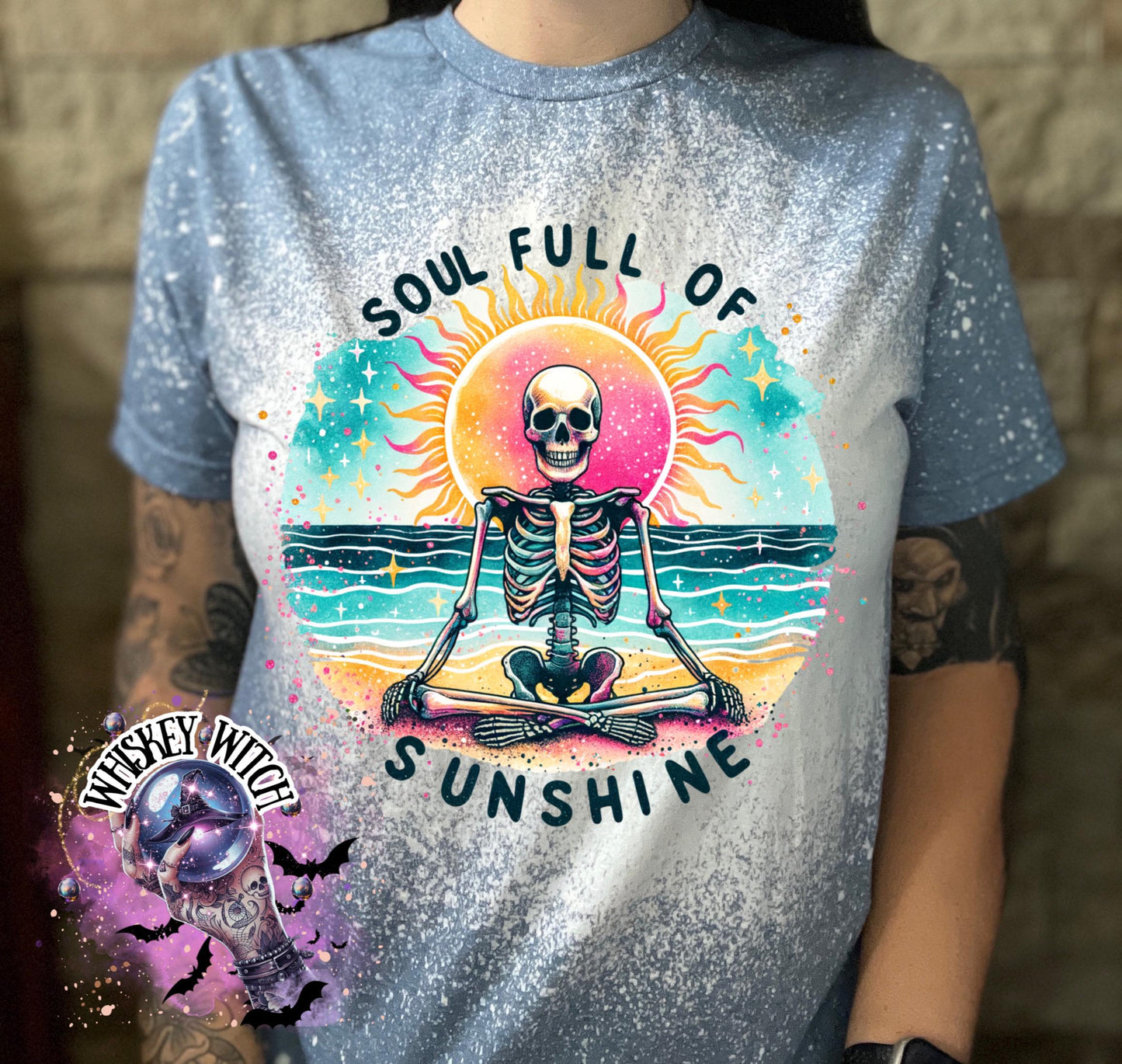 Light blue t-shirt with a bleached effect, featuring a colorful graphic of a skeleton meditating on a beach with a sun and the text "SOUL FULL OF SUNSHINE". The design includes turquoise, pink, yellow, and black elements.