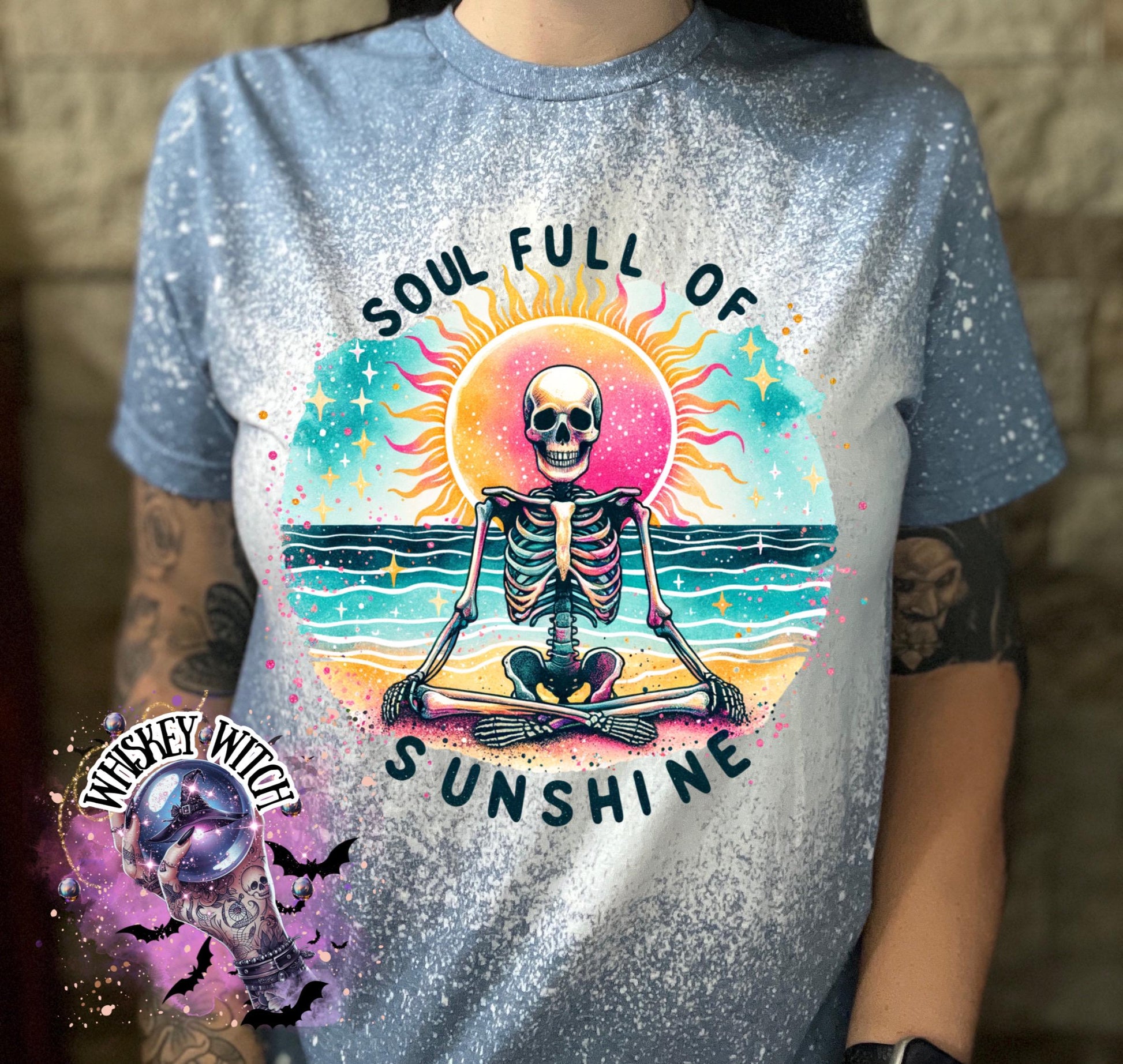 Light blue t-shirt with a bleached effect, featuring a colorful graphic of a skeleton meditating on a beach with a sun and the text "SOUL FULL OF SUNSHINE". The design includes turquoise, pink, yellow, and black elements.