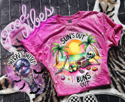 A pink t-shirt with a graphic of a skeleton relaxing on a beach with palm trees and the text "Sun's Out Buns Out." The shirt has a bleached effect.