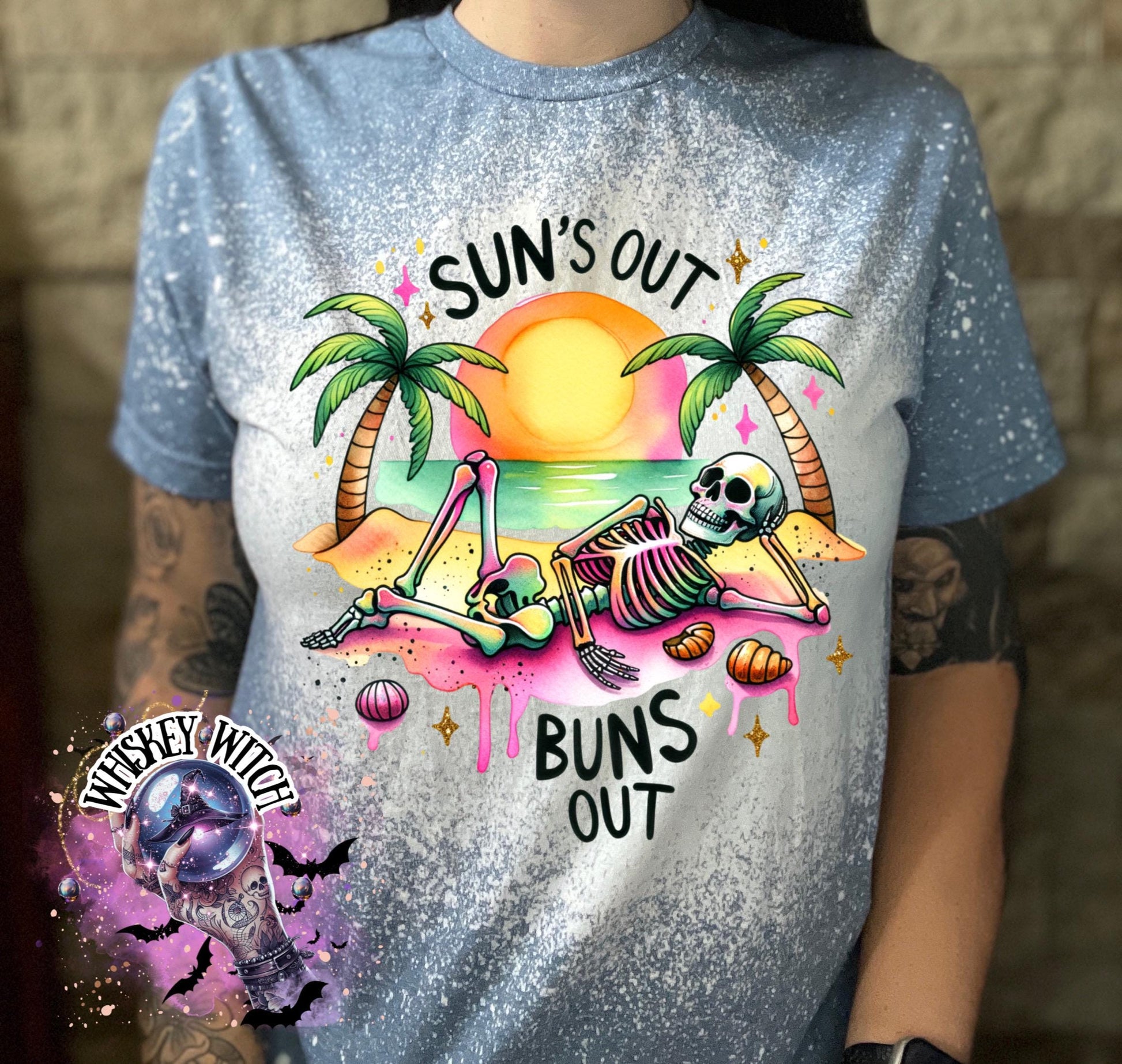 Light blue t-shirt with a bleached effect, featuring a skeleton relaxing on a beach with palm trees and a sunset. The text "SUN'S OUT BUNS OUT" is printed on the shirt.