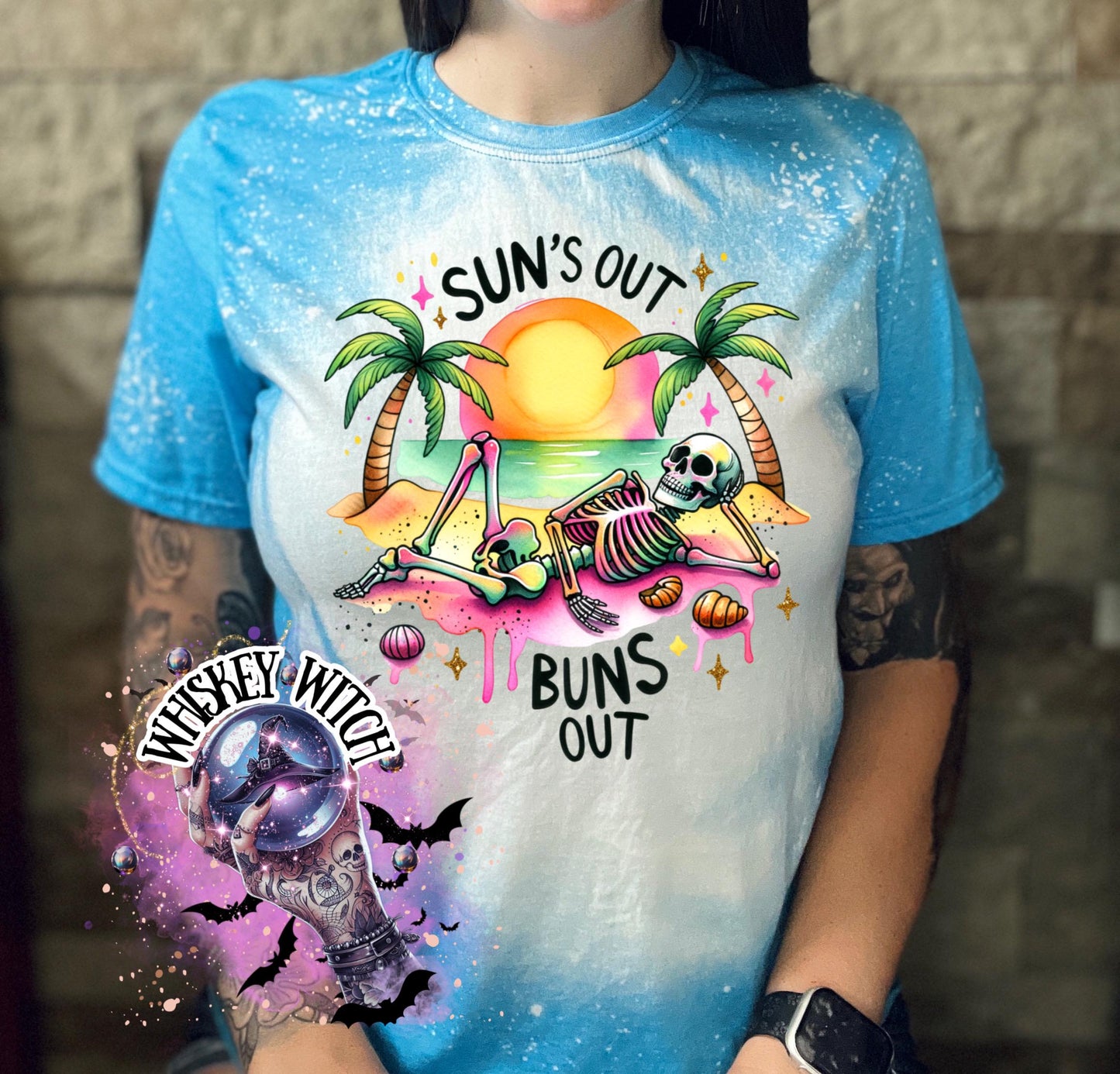A sapphire blue  t-shirt with a graphic of a skeleton relaxing on a beach with palm trees and the text "Sun's Out Buns Out." The shirt has a bleached effect.