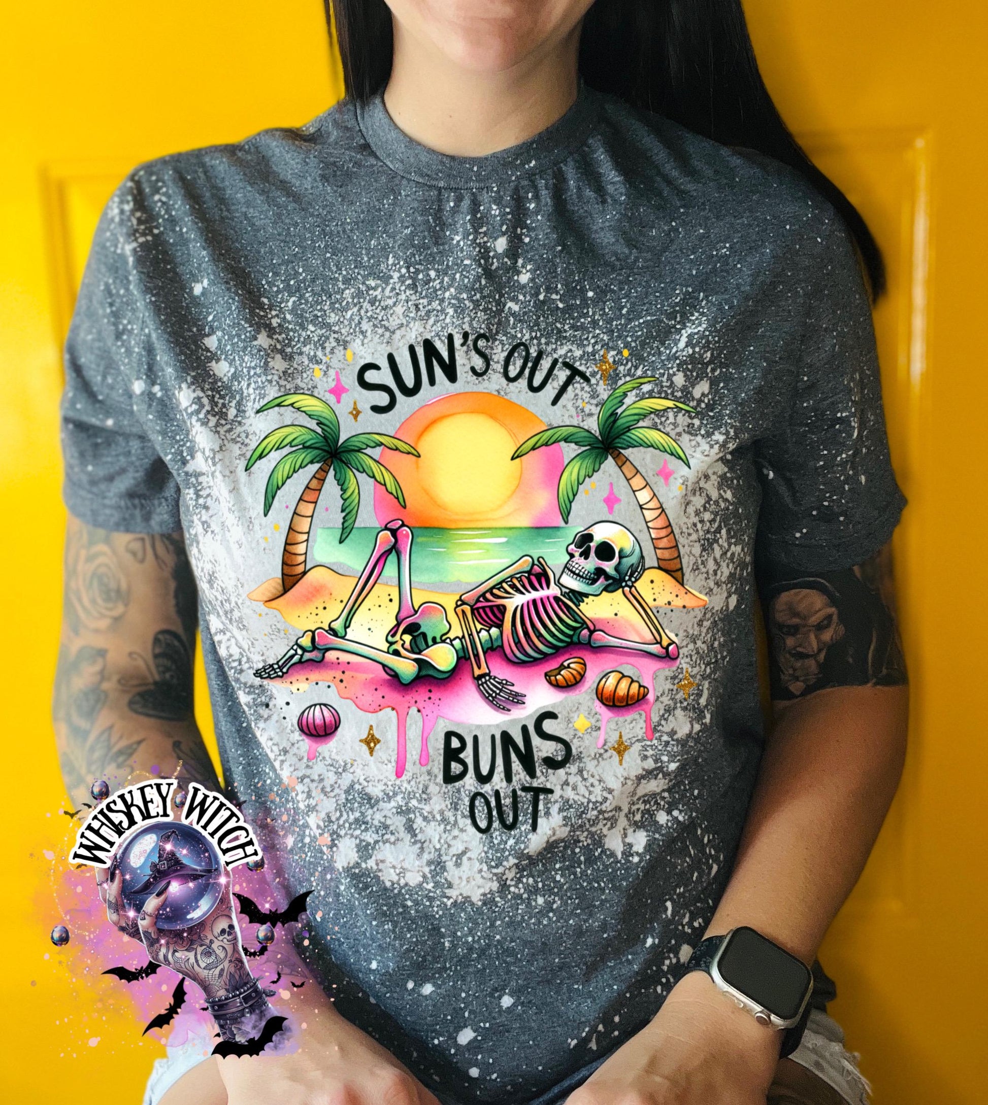 A grey t-shirt with a graphic of a skeleton relaxing on a beach with palm trees and the text "Sun's Out Buns Out." The shirt has a bleached effect.