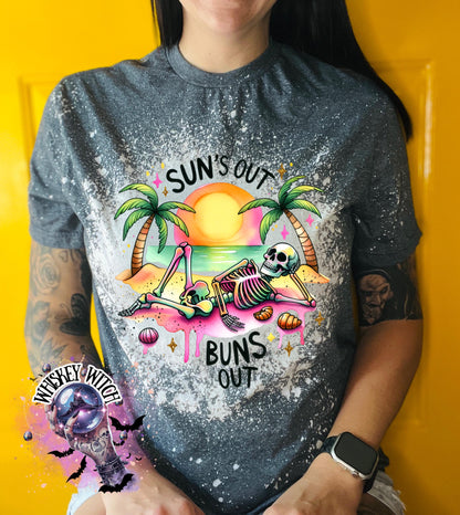 A grey t-shirt with a graphic of a skeleton relaxing on a beach with palm trees and the text "Sun's Out Buns Out." The shirt has a bleached effect.