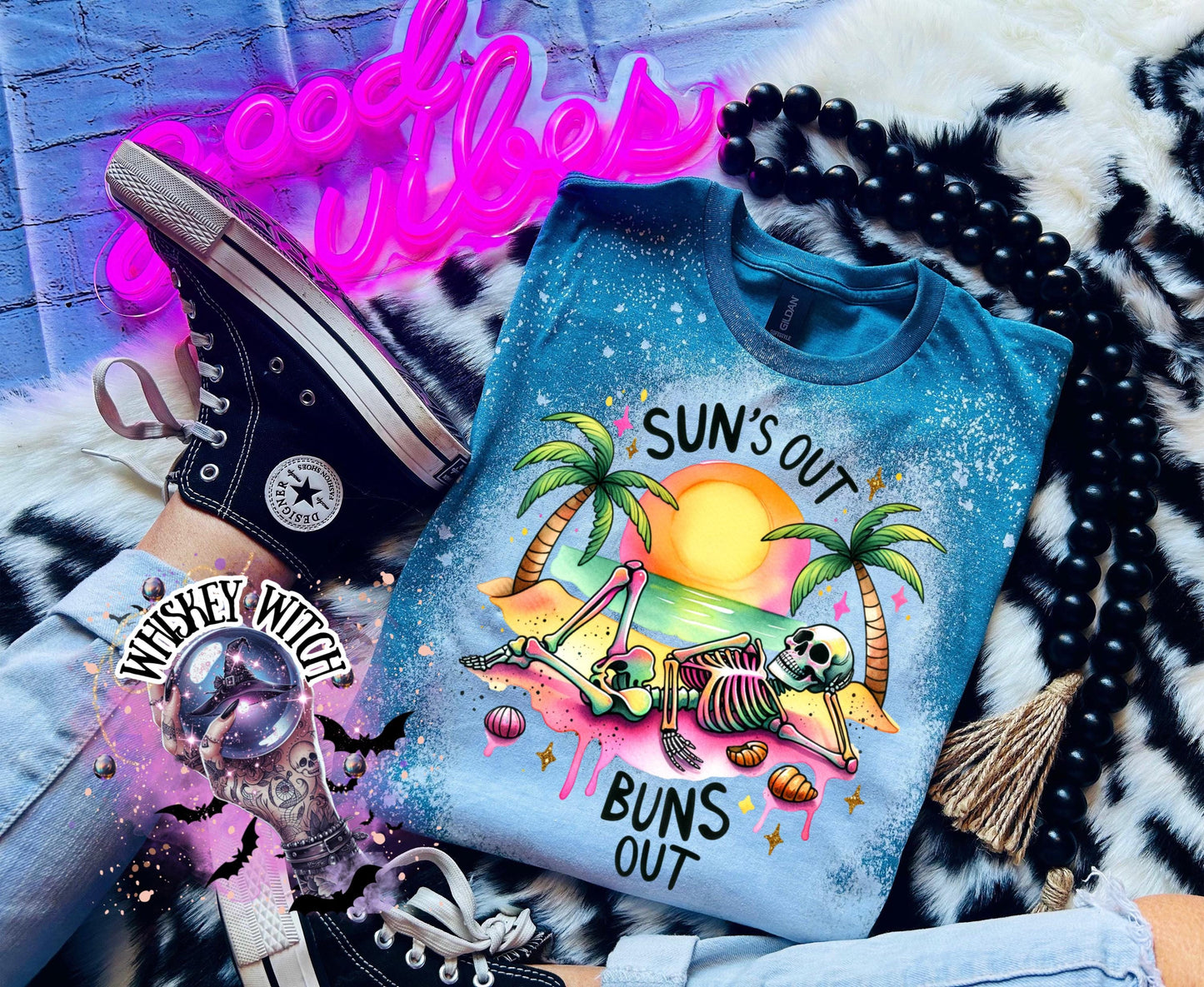 A Galapagos blue t-shirt with a graphic of a skeleton relaxing on a beach with palm trees and the text "Sun's Out Buns Out." The shirt has a bleached effect.