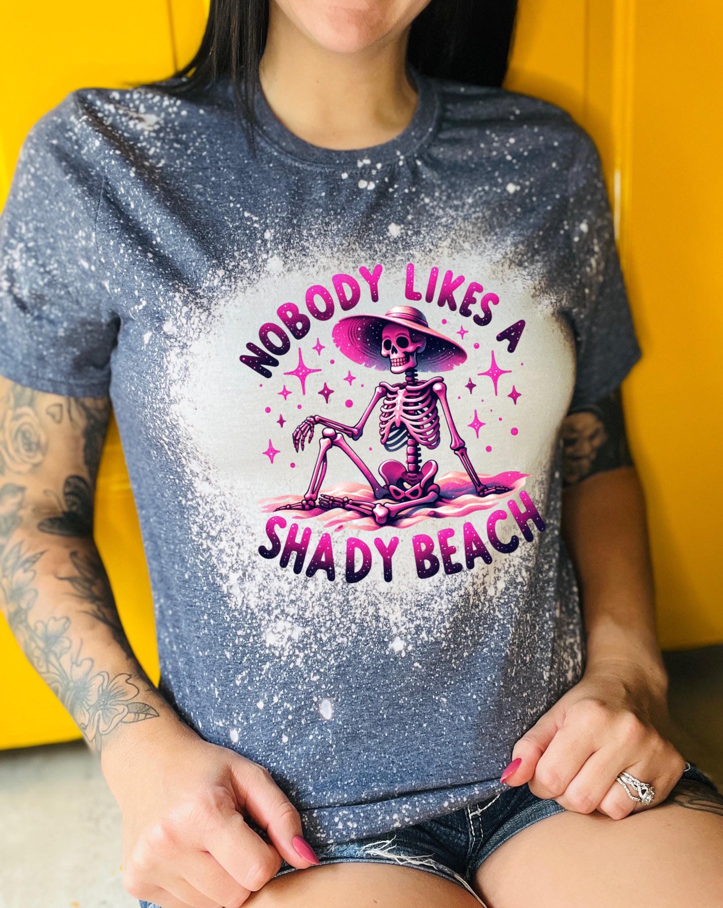 A navy blue  t-shirt with a bleached design featuring a skeleton wearing a hat, the text "Nobody Likes a Shady Beach," and star graphics. The shirt is paired with black and white checkered sneakers.